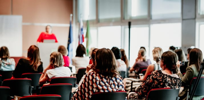 Business and entrepreneurship symposium. Female speaker giving a talk at business meeting. Audience in conference hall. Rear view of unrecognized participant in audience.