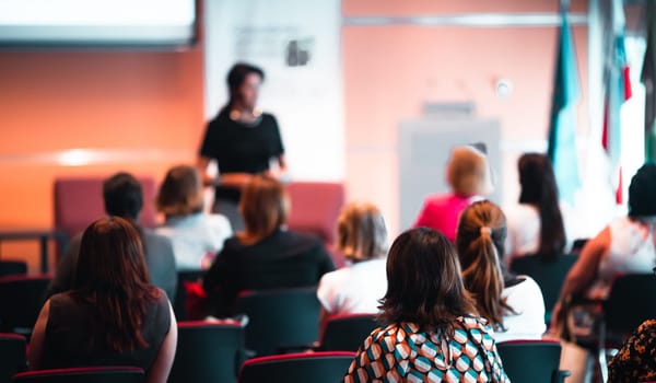 Business and entrepreneurship symposium. Female speaker giving a talk at business meeting. Audience in conference hall. Rear view of unrecognized participant in audience.