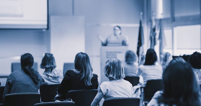 Business and entrepreneurship symposium. Female speaker giving a talk at business meeting. Audience in conference hall. Rear view of unrecognized participant in audience.