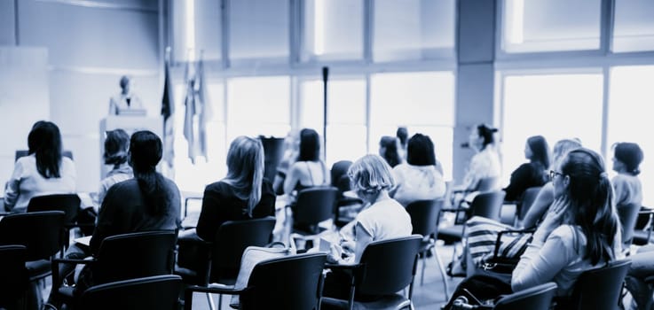 Business and entrepreneurship symposium. Female speaker giving a talk at business meeting. Audience in conference hall. Rear view of unrecognized participant in audience.
