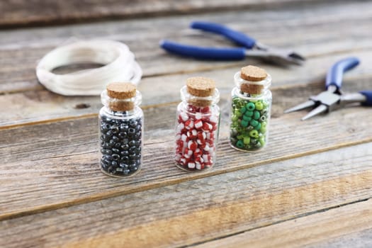 Red, green, and black seed beads in small jars on wooden surface with wire cutters, pliers and cord in background.
