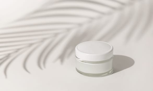 Cream jar with a blank white lid on white close up, Palm leaf hard shadow. Cosmetic packaging Mockup. Exotic natural cosmetics, monochrome composition, everyday skincare routine..