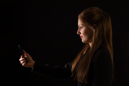 Young Caucasian woman holding smartphone in front of her face with black background. Side view portrait of female office worker interacting with modern technology at work.