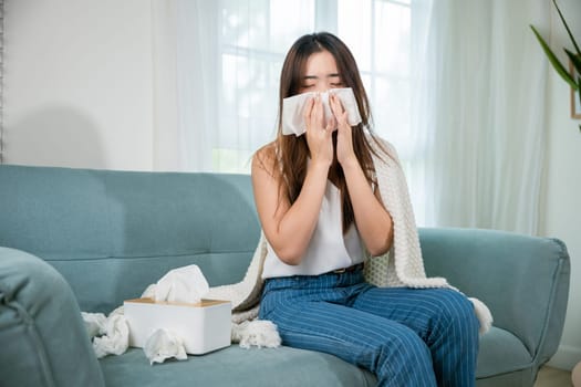Ill Asian young woman cold covered with blanket sitting on sofa and sneeze with tissue paper at home, Female sick allergic blowing nose sneezing in tissue at home, Flu health care