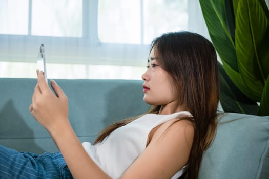 Happy Asian woman playing smart tablet at home while relaxing on sofa living room, Beautiful female sitting on couch using digital tablet pc, technology social media