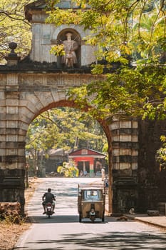 Old Goa, India. Auto Rickshaw Or Tuk-tuk Moving Through The Old Viceroy’s Arch. Famous Vasco Da Gama Gate Landmark And Historical Heritage.Street In Sunny Day.