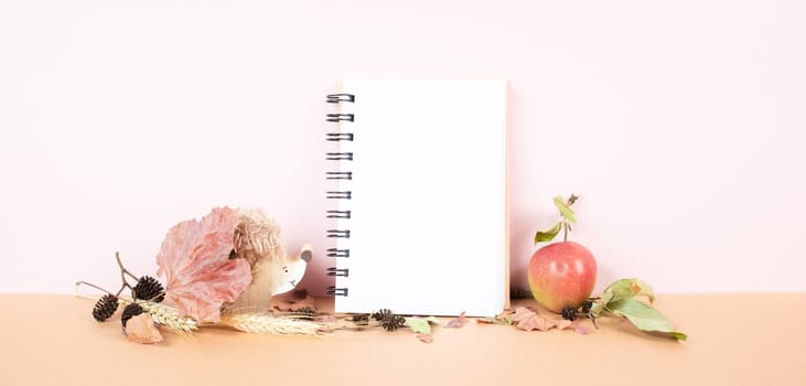 Blank notebook with an apple and a hedgehog, fall season background with colored leaves, office desk, pastel colors