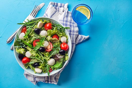 Green Salad with mozzarella cheese, black olives, arugula and tomato on blue table background
