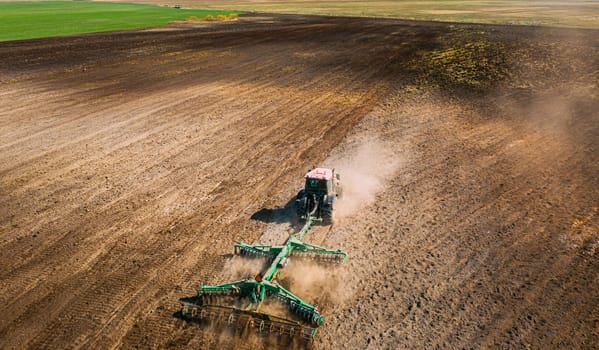 Aerial View. Tractor Plowing Field. Beginning Of Agricultural Spring Season. Cultivator Pulled By A Tractor In Countryside Rural Field Landscape. Dust Rises From Under Plows.