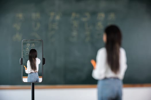 Back to school online class. Asian teacher teach on board while have blackboard lecture online through smartphone in classroom, woman standing front of class live video call online smart mobile phone