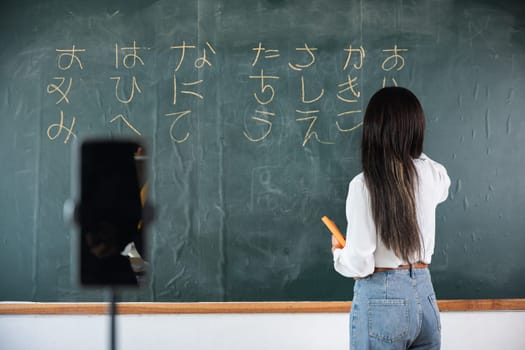 Back to school online class. Asian teacher teach on board while have blackboard lecture online through smartphone in classroom, woman standing front of class live video call online smart mobile phone