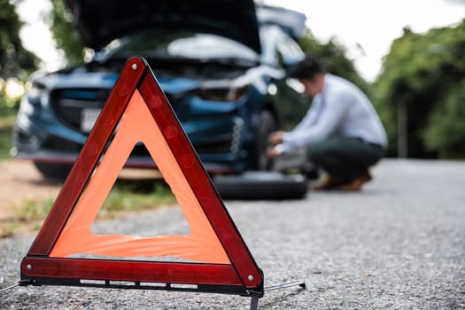 Asian businessman car broken has problems with wheel of his car during go to work in morning he changing replace rubber tire on wheels, business man have warning sign on road, problem transportation