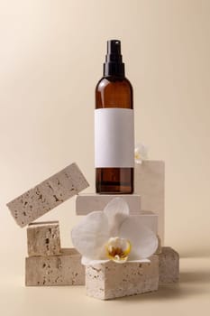 Dark glass cosmetic bottle with blank label near white orchid flowers and stones on light yellow, close up, mockup. Skincare handmade beauty product, tonic or lotion. Exotic natural cosmetics..