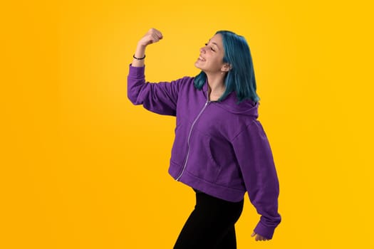 Cute young gilr with blue hair doing winner gesture, celebrate clenching fists. Isolated on plain yellow background studio portrait.