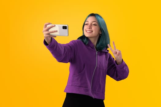 Cheerful nice young girl with blue hair taking selfie on cellphone and smiling. Isolated on yellow baclground