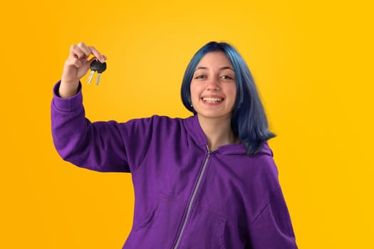 Smiling young woman student with blue hair holding keys over yellow background