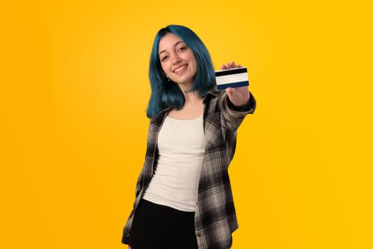 Smiling young woman student with blue hair holding a credit card over yellow background