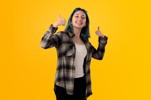 Smiling young woman student with blue hair doing positive gestures with her hands isolated over yellow background