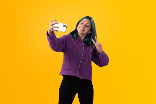 Cheerful nice young girl with blue hair taking selfie on cellphone and smiling. Isolated on yellow baclground