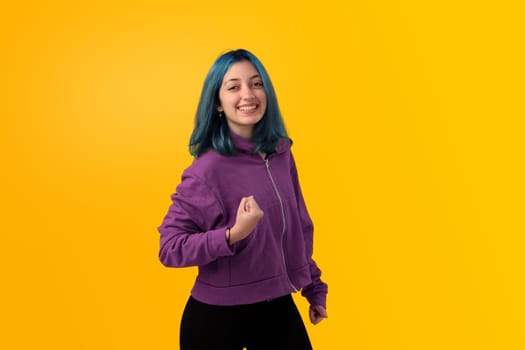 Cute young gilr with blue hair doing winner gesture, celebrate clenching fists. Isolated on plain yellow background studio portrait.