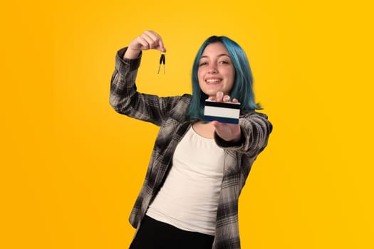 Smiling young woman student with blue hair holds a credit card and key over a yellow background