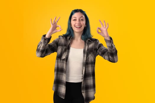 Smiling young woman student with blue hair doing positive gestures with her hands isolated over yellow background