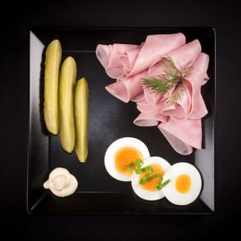Elegant cold appetizer, ham, egg and pickle on black plate on black background in gourmet restaurant.