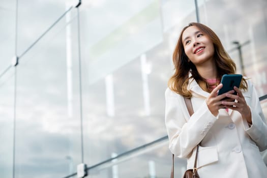 Asian businesswoman working with mobile phone standing against street front building near office, Portrait successful business woman smiling holding smartphone use application chat online in morning