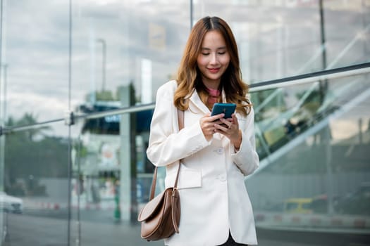 Asian businesswoman working with mobile phone standing against street front building near office, Portrait successful business woman smiling holding smartphone use application chat online in morning