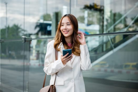 Asian businesswoman working with mobile phone standing against street front building near office, Portrait successful business woman smiling holding smartphone use application chat online in morning