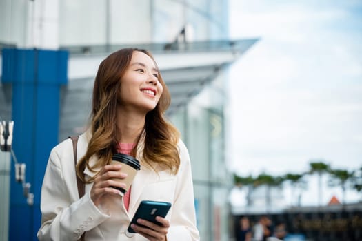 Young business woman smiling holding mobile phone with coffee take away going to work early in morning, Asian businesswoman with smartphone and cup coffee standing against street building near office
