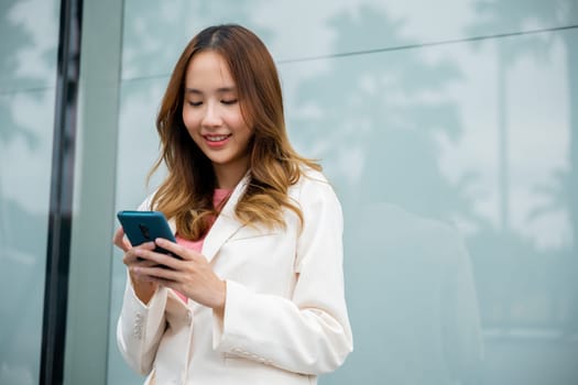 Asian businesswoman working with mobile phone standing against street front building near office, Portrait successful business woman smiling holding smartphone use application chat online in morning