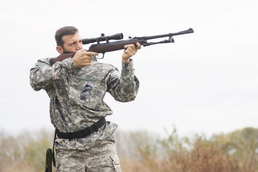 The hunter in military uniform is aiming and shooting a weapon outdoors, hunting sport.