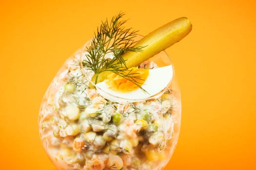 Decorated Russian salad with egg, pickle and dill is in drinking glass on orange background.