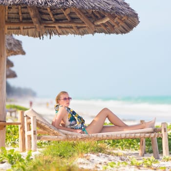 Women sunbathing on dack chair under wooden umbrella on stunning tropical beach. Kiteboarders turquoise blue lagoon of Paje beach, Zanzibar, Tanzania in the background.
