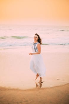 Goa, India. Young Caucasian Woman In White Dress Walking Along Seashore, Looking At Camera And Smiling In Summer Sunlight.