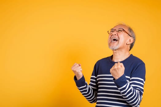 Portrait smiling senior old man lifestyle he arms raised to celebrating his success, Happy Asian elder man with glasses excited say yes gesture like winner studio shot isolated on yellow background