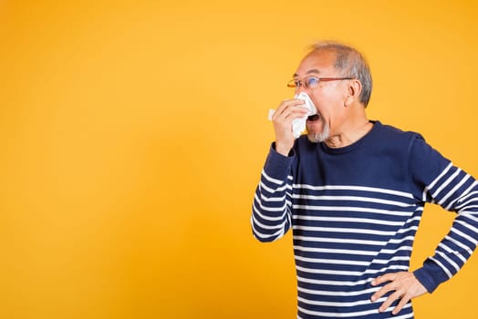 Asian elder man cold having flu and sneezing from sickness virus problem use tissue studio shot isolated on yellow background, Portrait senior old man blowing nose with tissue, pensioner unwell