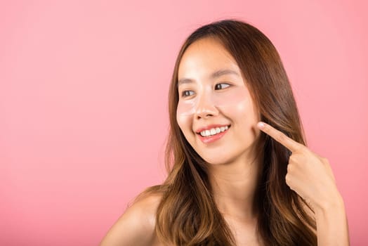Eye treatment. Asian beautiful young woman smiling point finger to cosmetic patches gel under eyes, Female with natural makeup hydrogel eye patch on face, isolated on pink background, skincare