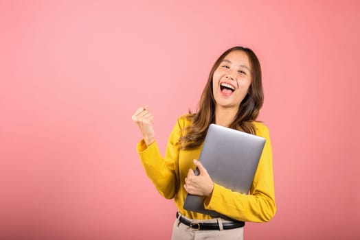Portrait of happy Asian beautiful young woman confident smiling face holding laptop computer raise hand and say yes! excited celebrating success, studio shot isolated on pink background, copy space