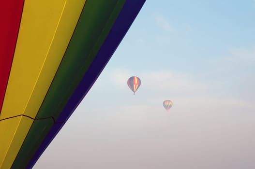 Two hot air balloons can be seen high up in the sky and partially obscured by the fog in the air, The side of another balloon fills the side of the image up close.