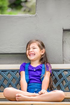 A pretty little girl,with long dark hair, and purple shirt, sits on an outside bench in the garden with her legs folded, while laughing with joy.