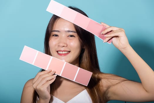 Portrait of happy female hold color card under sun protection studio shot isolated on blue background, Asian beautiful young woman smiling and holding skin color scale paper, skincare beauty face