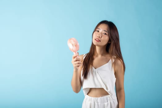 Asian beauty young woman holding pink portable electric mini fan near her face studio shot isolated on blue background, Female hand hold small plastic fan handheld she enjoying cool wind blowing