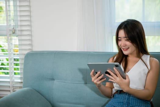 Happy Asian woman playing smart tablet at home while relaxing on sofa living room, Beautiful female sitting on couch using digital tablet pc, technology social media