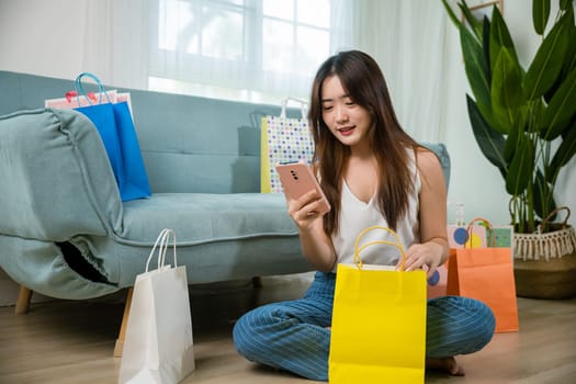 Excited Asian buying woman holding mobile phone with many shopping bags at home in living room shopping, Happy female online shopping payment sitting on couch with smartphone, Black Friday