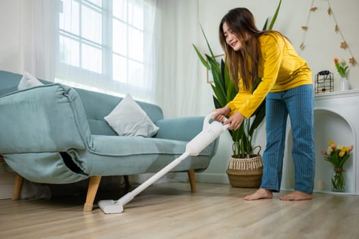 Housewife female dust cleaning floor under sofa or couch furniture with vacuum cleaner, Happy Asian young woman with accumulator vacuum cleaner at home in living room, household and housework concept