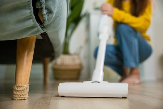 Happy Asian young woman with accumulator vacuum cleaner at home in living room, housewife female dust cleaning floor under sofa or furniture with vacuum cleaner, household and housework concept
