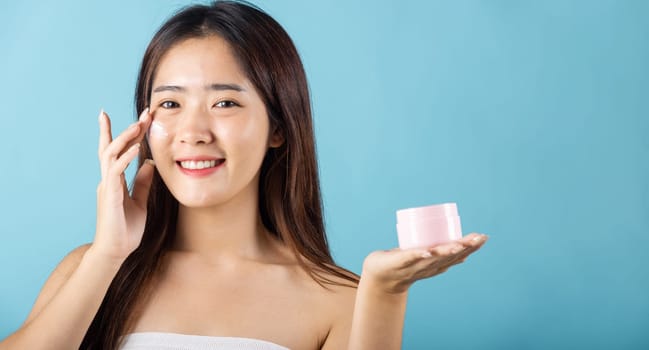 Asian young woman are not very beautiful hold jar of skin cream for face and apply anti-aging moisturizer studio isolated on blue background, Happy female pointing finger her face, pursuit of beauty