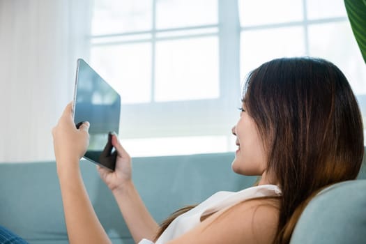Happy Asian woman playing smart tablet at home while relaxing on sofa living room, Beautiful female sitting on couch using digital tablet pc, technology social media